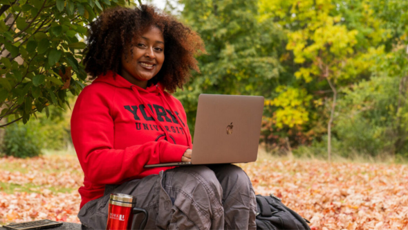 A Black female student wearing a red York hoodie sits with her laptop and York tumbler among fall leaves. 